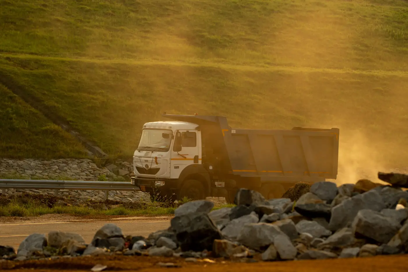 A dump truck driving along a dusty road past piled rock at dusk.